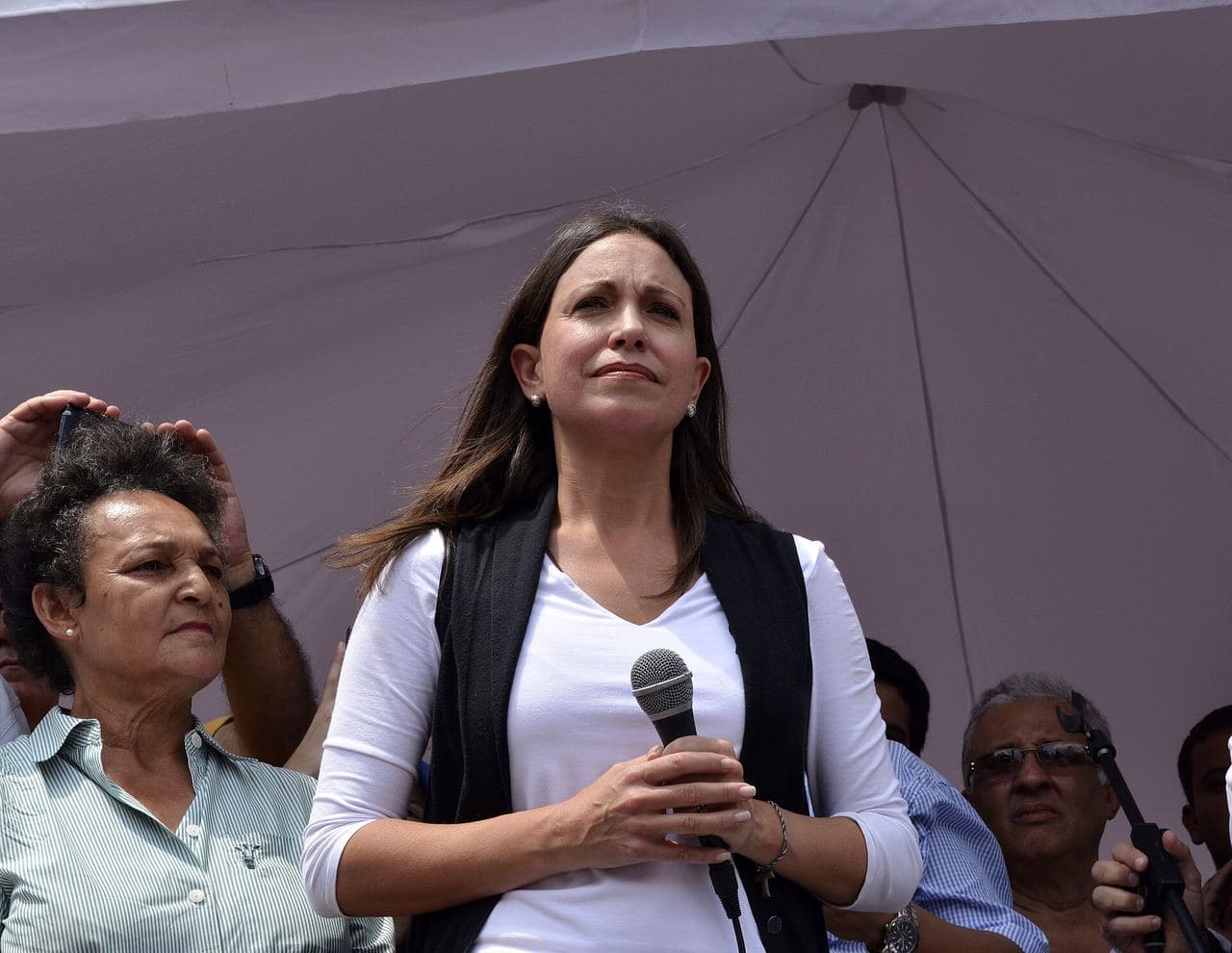 María Corina Machado, dressed in white and holding a microphone, standing under a canopy among other people.
