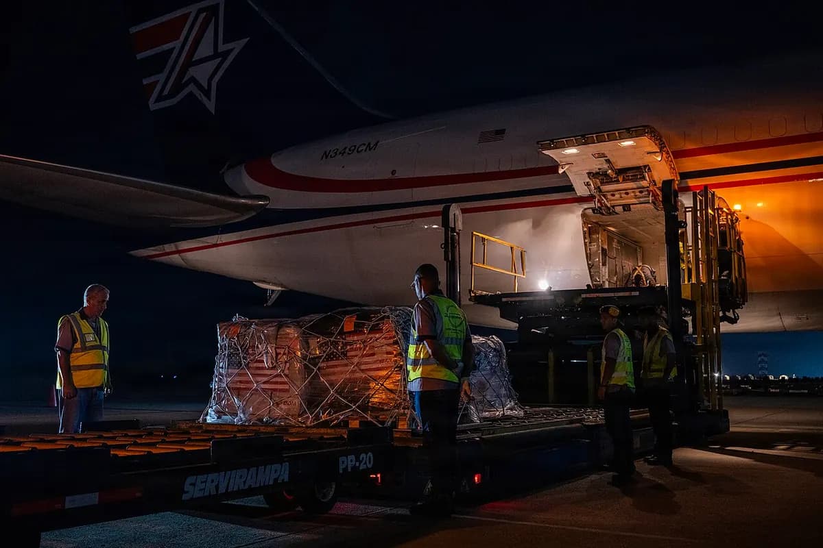 At night, beside the open cargo door of a plane, workers in reflective vests unload pallets of net-wrapped boxes onto a ramp.