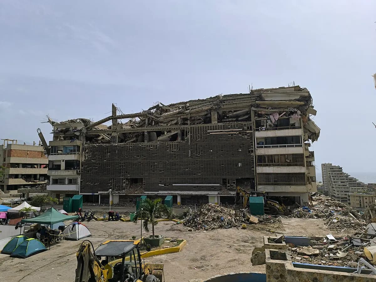A long building with its top floors caved in and its facade torn apart; in front, an open lot with camping tents, piles of rubble and a backhoe.