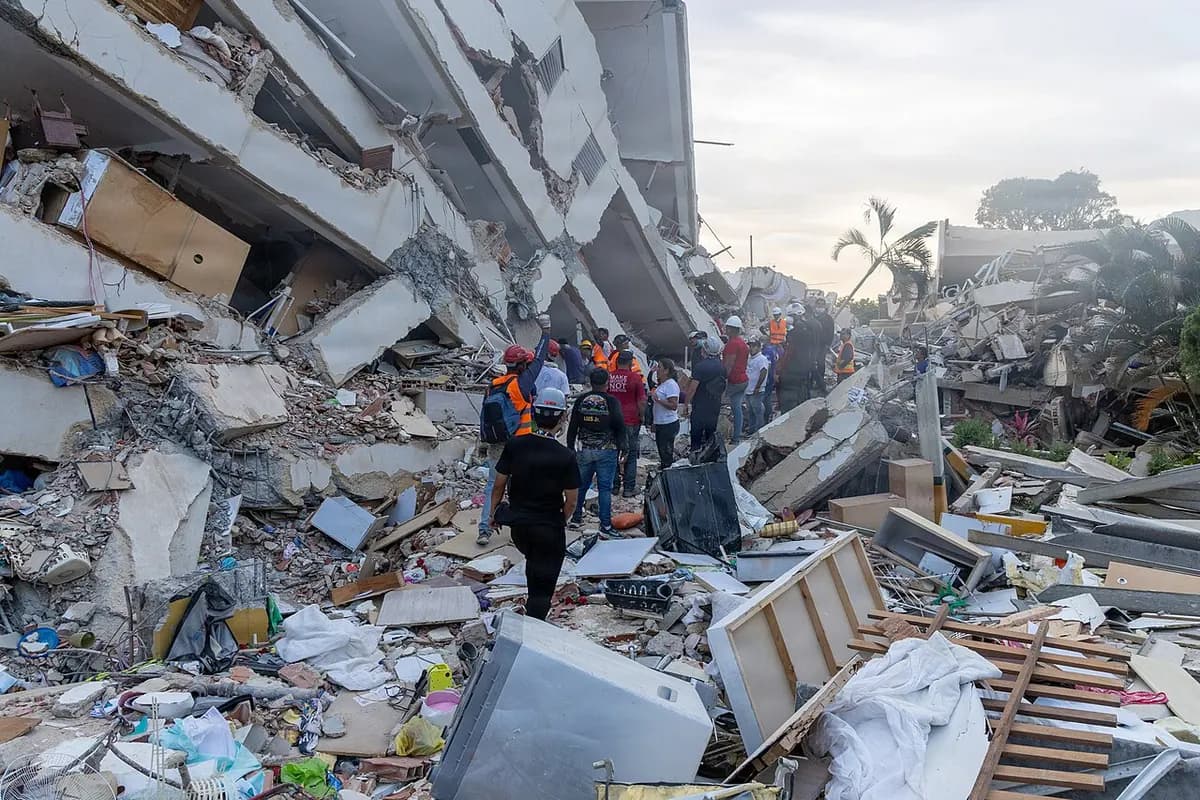 A group of people, seen from behind and at a distance, walks through rubble, furniture and appliances beside a tilted, broken building.