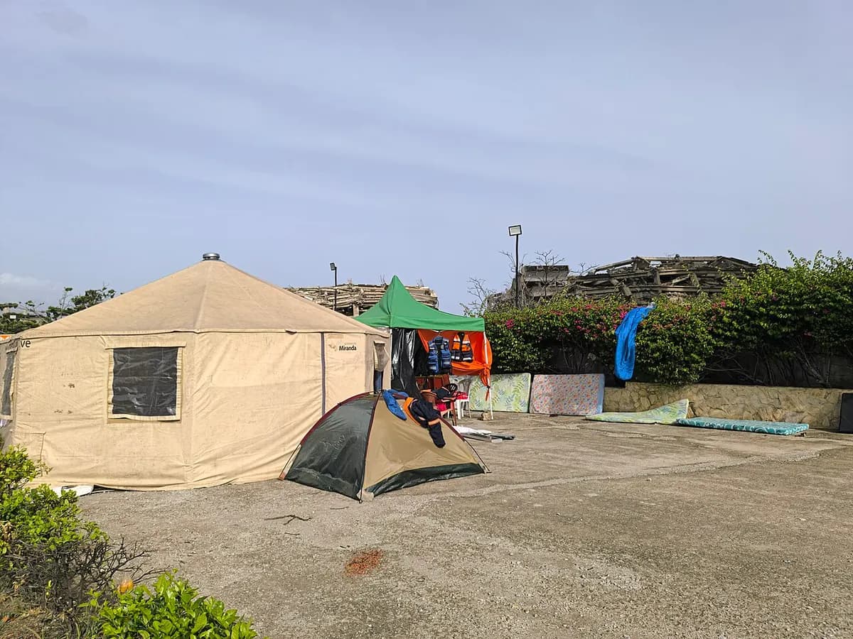 A large canvas tent, a small camping tent and a green canopy on a concrete lot, with mattresses propped against a wall; in the background, the remains of collapsed buildings.