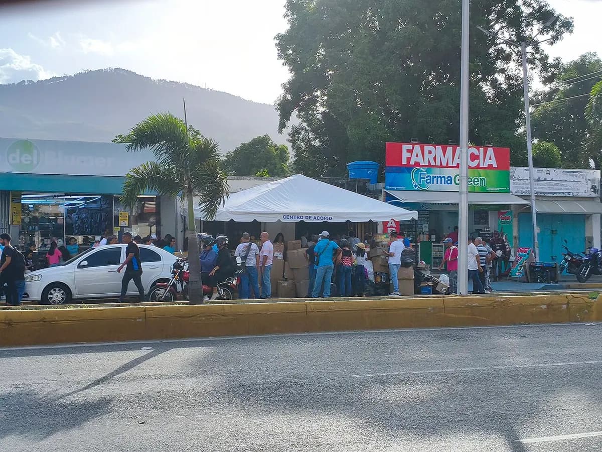 A white canopy with a “Centro de acopio” (collection centre) sign on an avenue sidewalk, with stacked boxes and people lining up to leave donations, in front of a pharmacy.