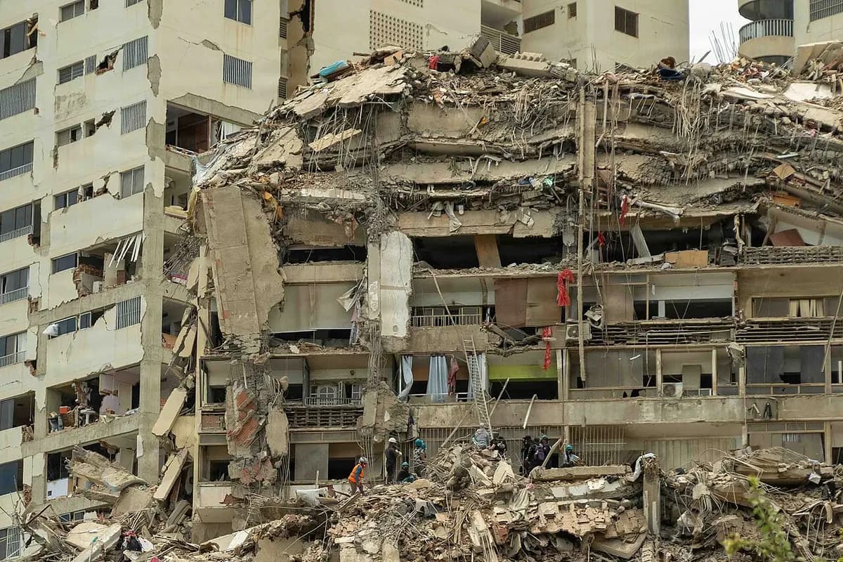 An apartment block whose upper floors are crushed one on top of another; below, on the mound of rubble, several small figures in the distance search through the debris.