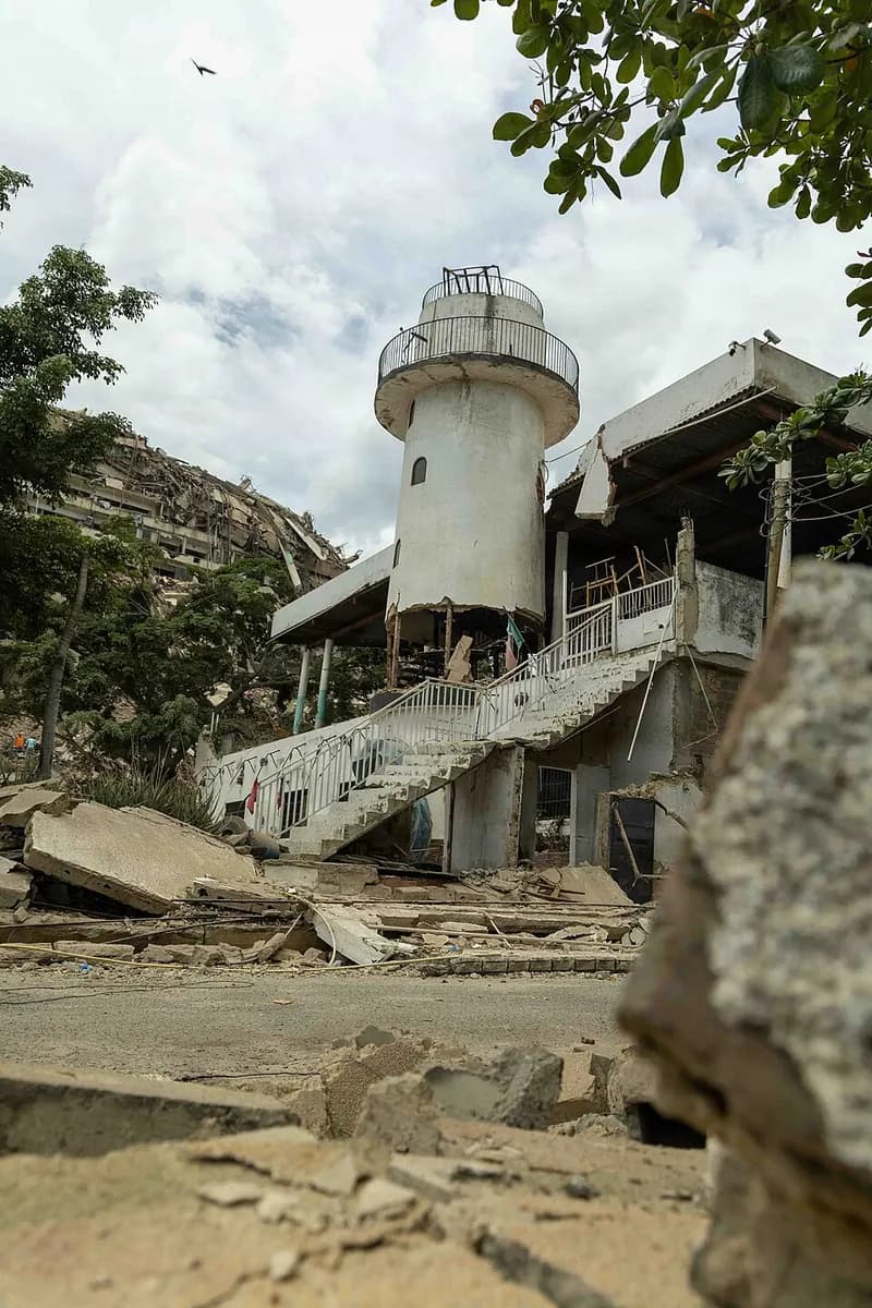 A venue with a white lighthouse-shaped tower, its outdoor staircase and roof damaged, and concrete blocks in the foreground; behind it, on the left, a collapsed building.