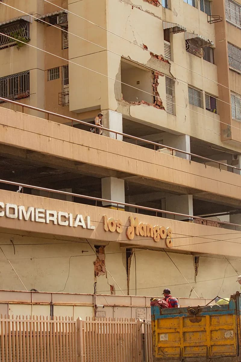 A residential building with a cracked facade: on one floor an apartment's outer wall is missing and the broken bricks show; below, vertical cracks above the shopping-centre sign.