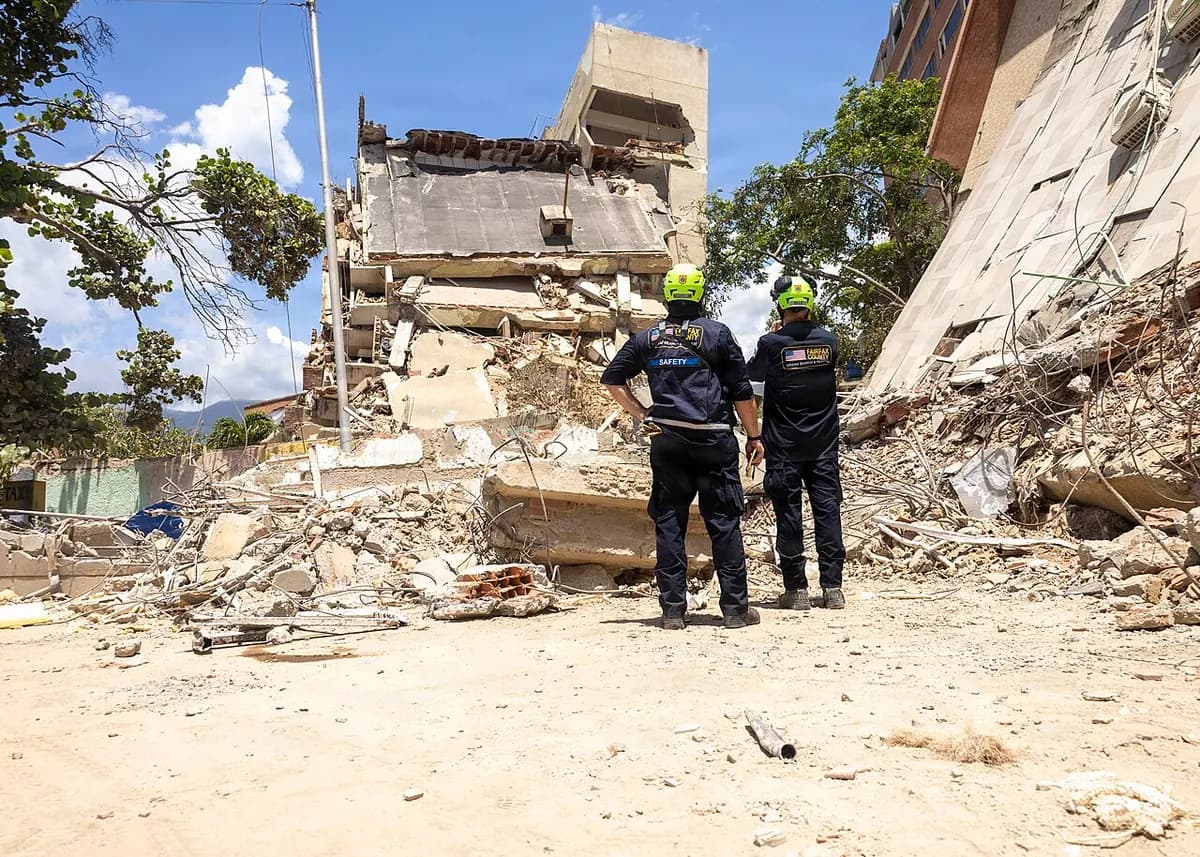 Two rescuers in yellow helmets, seen from behind, look at a multi-storey building collapsed onto the street; on the right, the wall of another building has fallen diagonally onto the rubble.