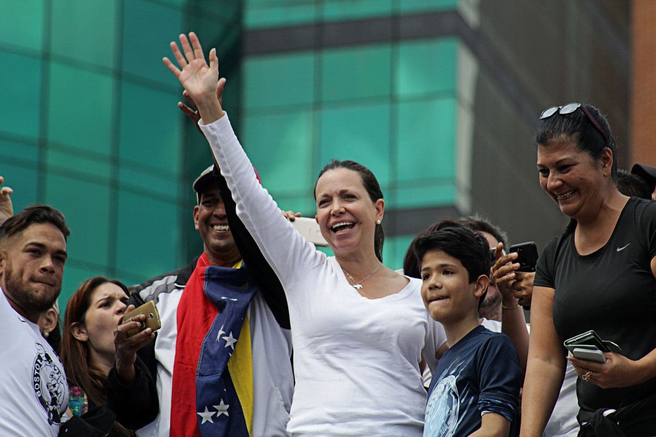 María Corina Machado smiles and raises her arm to wave, surrounded by protesters; one of them carries a Venezuelan flag.