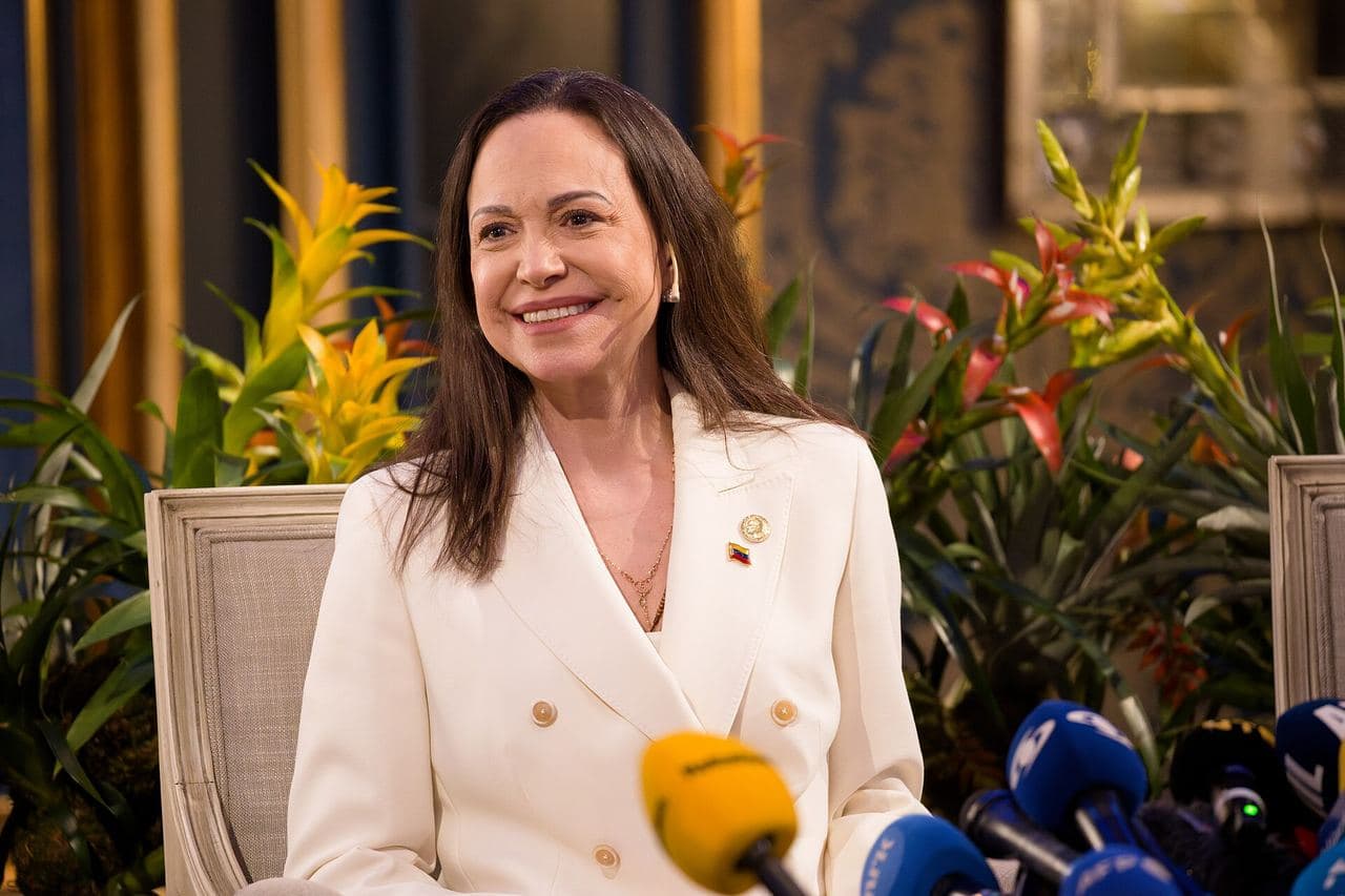 María Corina Machado, in a white jacket, smiles while seated in front of several microphones at a press conference.